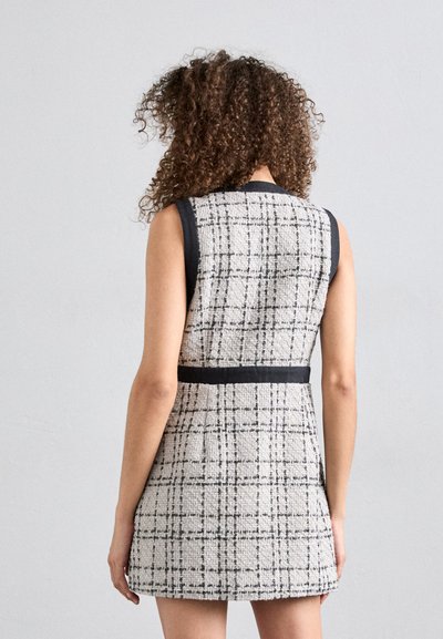 Robe sans manches en tissu beige texturé, avec des accents noirs et une taille ceinturée. Le design présente un motif à carreaux.