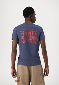 Tamsiai mėlynas marškinėliai su raudonu tekstu nugaroje, kuriame parašyta "The Replay Blue Jeans" su retro dizainu. Minkšta medžiaga, trumpomis rankovėmis.