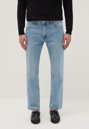 Mann trägt hellblaue Jeans mit geradem Schnitt, ein schwarzes langärmeliges Oberteil und schwarze Schnürschuhe, steht vor einem schlichten Hintergrund.