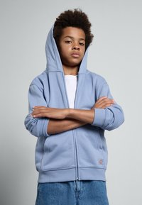 Moletão azul claro com fecho e textura, com bolsos tipo Hoodie. Usado sobre uma t-shirt branca com um logótipo perto da bainha.