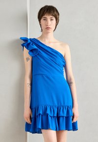 Robe bleue à une épaule, en tissu lisse, avec un décolleté à volants et un ourlet à volants superposés, dotée d'un corsage ajusté et d'une coupe ample.