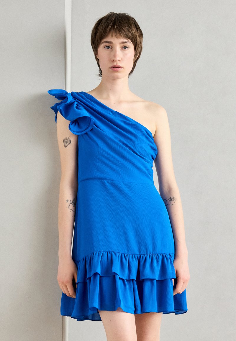 Robe bleue à une épaule, en tissu lisse, avec un décolleté à volants et un ourlet à volants superposés, dotée d'un corsage ajusté et d'une coupe ample.