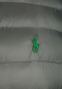 Grå puffjacka i närbild med en grön broderad hästlogotyp, med quiltat mönster och sydda detaljer.