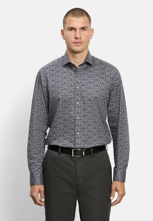 Uomo che indossa una camicia a maniche lunghe con motivo a foglie, pantaloni scuri e cintura nera, rivolto in avanti su uno sfondo semplice.