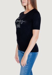 Maglione nero a maniche corte con scollo rotondo e decorazioni in cristallo che spelling "Guess est 1981" sul davanti.