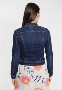 Chaqueta de denim en azul oscuro, con un diseño ajustado, puños abotonados y detalle en el yugo trasero. Llevada sobre una falda de estampado floral.