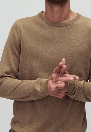 Person in einem braunen Pullover, die Hände vor der Brust verschränkt, vor einem schlichten Hintergrund.