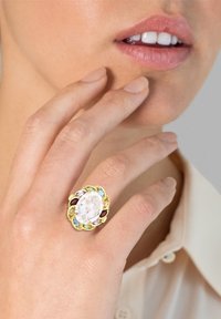 Bague en or avec une pierre ovale transparente, entourée de gemstones colorés dans des teintes de bleu, rouge et vert ; un doigt touchant doucement le menton.