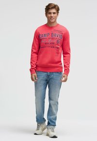 Rotes Sweatshirt mit grafischem Text und Logo in Dunkelblau, kombiniert mit hellblauen Jeans und weißen Sneakers mit grauen Akzenten.