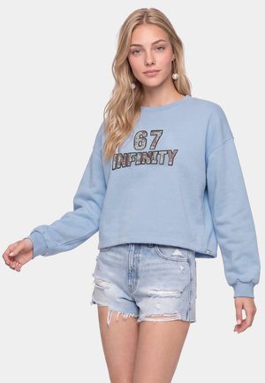 Kvinde med langt blondt hår iført perleøreringe, en lyseblå sweatshirt med teksten "67 INFINITY", og revnede denimshorts.