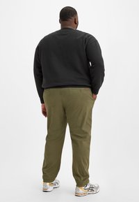 Schwarzer Rundhalsausschnitt-Pullover, olivgrüne Hosen und silberne Sportschuhe mit gelben Akzenten. Sichtbare Gesäßtaschen an den Hosen.