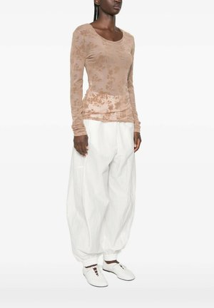 Vrouw draagt een lichtbruine top met lange mouwen en bloemenpatroon, losse witte broek samengebonden bij de enkels en witte veterschoenen.