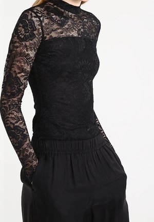 Femme portant un haut en dentelle noire à manches longues, col montant et manches transparentes, associé à un pantalon noir ample, mains dans les poches.