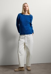Suéter azul de punto con escote amplio, pantalones blancos holgados y zapatos beige. El suéter presenta una tela texturizada y puños acanalados.