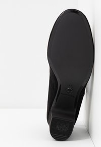 Semelle d'une chaussure noire avec une semelle lisse, petit talon et texte « ANTI SLIDE » sur le talon, taille 37, debout contre une surface blanche.