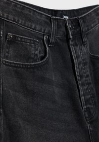 Jean en denim noir avec taille haute, fermeture à boutons et design classique à cinq poches, avec coutures et textures contrastées.