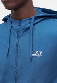 Homme portant un sweat à capuche zippé bleu EA7 Emporio Armani avec cordons, montré du cou à la poitrine sur fond blanc.