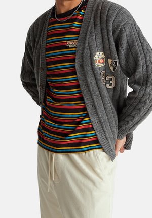 Homme portant un cardigan gris en maille avec des patchs sur un t-shirt rayé noir, rouge, bleu et jaune, associé à un pantalon beige clair.