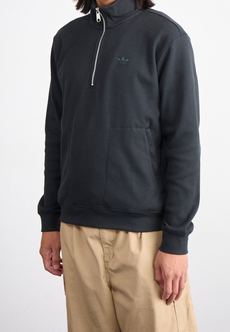 Sudadera de media cremallera en gris oscuro con textura acanalada, con cuello con cremallera y un bolsillo delantero. Incluye un logo sutil en el pecho.