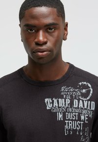 Schwarzer Sweatshirt mit rundem Ausschnitt, der weiße grafische Texte und Akzente aufweist. Der Stoff wirkt strukturiert mit einem subtilen Glanz.