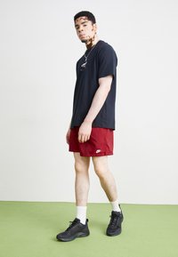 Fekete póló, piros nadrág fehér Nike logóval, fekete sportcipő, fehér zoknik, zöld padlón állva, egyszínű háttér előtt.
