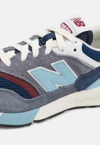 Graue und blaue Sneakers mit strukturiertem Wildleder- und Mesh-Obermaterial, roten Akzenten, einem hellblauen Lederlogo und weißen Schnürsenkeln.