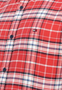 Camicetta a quadri rossi con motivi a scacchi blu e bianchi, caratterizzata da bottoni scuri e un piccolo logo del marchio sulla parte sinistra del petto.