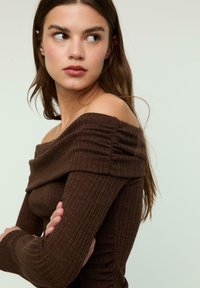 Mlada ženska z dolgimi rjavimi lasmi, oblečena v rebrast rjav off-shoulder top, gleda vstran s prekrižanima rokama proti nevtralni ozadju.