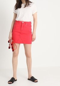 Jupe en denim rouge à coupe ajustée et poches avant, associée à un t-shirt beige clair et des sandales noires ouvertes. Accessoirisée avec un petit sac.