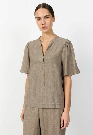 Frau trägt ein locker sitzendes, kurzärmeliges, beiges Leinenoberteil mit V-Ausschnitt und passenden weit geschnittenen Hosen vor einem einfarbigen weißen Hintergrund.