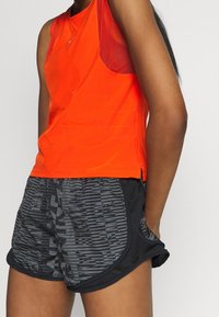 Pomarańczowy sportowy top na ramiączkach z siateczkowymi akcentami, w połączeniu z czarnymi szortami o teksturowanym wzorze. Oba elementy odzieży mają luźny krój.