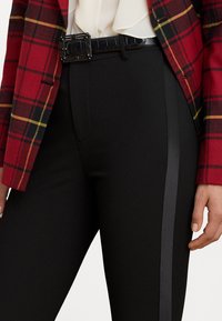 Crne prilagođene hlače s sjajnom bočnom trakicom, uparene s blejzerom u crvenoj i žutoj karo uzorku. Outfit sadrži dekorativni crni pojas.
