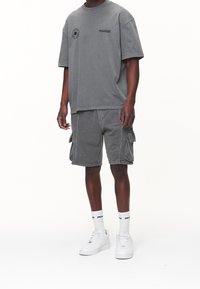 Hombre vistiendo una camiseta gris oversized y un conjunto de pantalones cargo, con calcetines blancos con logo y zapatillas blancas, de pie frente a un fondo blanco.