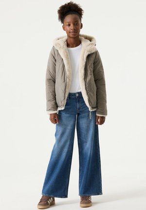 Meisje staand in een taupekleurige imitatie-shearling capuchonjas, wit shirt, wijde blauwe jeans en bruine sneakers tegen een effen achtergrond.