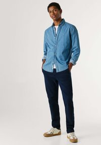 Light blue denim shirt with a button-up design, rolled sleeves, paired with navy chinos and white sneakers with tan accents.