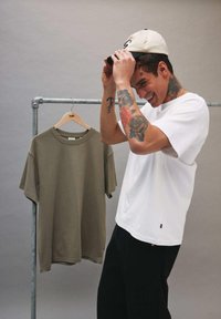 Jeune homme avec des tatouages ajustant une casquette blanche, debout à côté d'un t-shirt vert olive accroché à un portant métallique sur un fond uni.