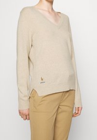 Beige v-ringad tröja i mjukt stickat tyg, med ribbade detaljer och en logotyp vid fållen. Kombinerad med ljusbruna byxor.