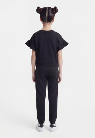 Παιδί που στέκεται με την πλάτη προς τα εμπρός, φορώντας μαύρο μπλουζάκι με κοντά μανίκια και βολάν, μαύρο παντελόνι, λευκά αθλητικά παπούτσια και διπλό κότσο στα μαλλιά.