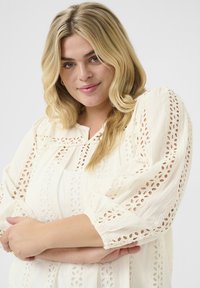 White blouse with intricate eyelet lace detailing on sleeves and bodice. Lightweight fabric, featuring a relaxed fit and round neckline.