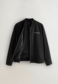 Fekete bomber dzseki ezüst cipzárral, részben nyitva, hálós bélést mutatva, és fehér, díszes betűs felirat a bal mellrészen.