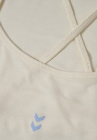 Cremefarbener Sportoberteil mit gekreuzten Trägern, aus glattem Material und einem kleinen blauen Logo-Detail unten links.