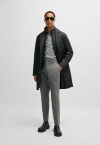Piumino nero sopra una camicia grigia con bottoni, abbinato a pantaloni grigi a fantasia e stivali neri chunky. Gli occhiali da sole completano il look.