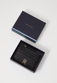 Black textured wallet with gold hardware inside a dark blue box. Features a zip closure and embossed pattern on surface.