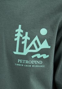 Forestgroene t-shirt met een lichtturquoise grafiek van bomen, bergen en water, met de tekst "PETROLIND TIMBER-CREW MILWAUKEE".