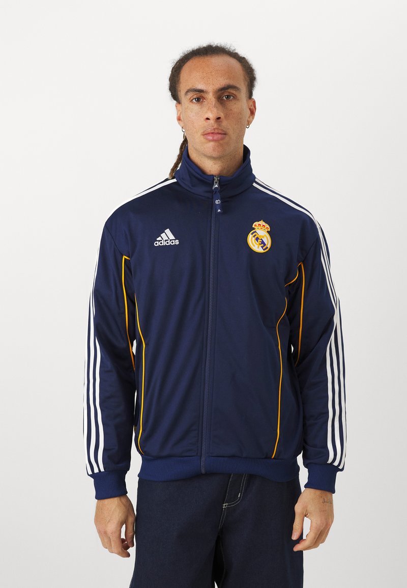 Veste de survêtement marine en tissu lisse, avec des rayures blanches sur les manches, des accents jaunes et un logo du Real Madrid sur la poitrine.