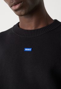 Pulover tricotat negru cu guler cu fâsle, având un patch brodat albastru cu logo-ul "HUGO" pe partea stângă.