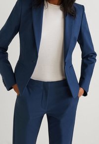 Veste de costume bleu cintrée avec revers crantés, assortie à un pantalon coordonné. Chemise blanche ajustée visible en dessous. Texture de tissu lisse.