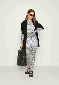 Cardigã longo preto sobre um bodysuit com estampa floral cinza e leggings. Uma tote preta oversized e sandálias de plataforma completam o visual.