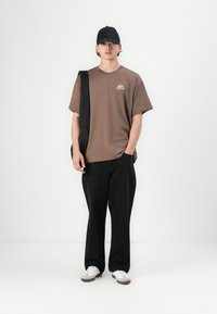 Hnědé bavlněné tričko s logem, černé kalhoty širokého střihu a bílé tenisky. Součástí outfitu jsou také černá čepice a texturovaná černá taška.