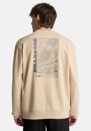 Mężczyzna w beżowej bluzie z mapą topograficzną i napisem „Billabong Adventure Division” na plecach, stoi tyłem na białym tle.